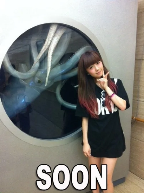 A young woman with long brown hair and a black t-shirt poses cheerfully in front of a large circular window, making a peace…