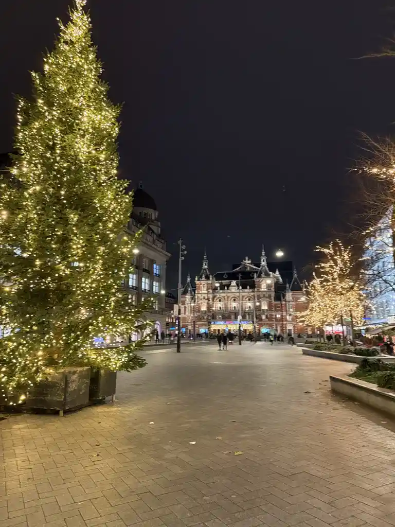 A nighttime city square scene featuring a large, brightly lit Christmas tree on the left, with ornate buildings and other…