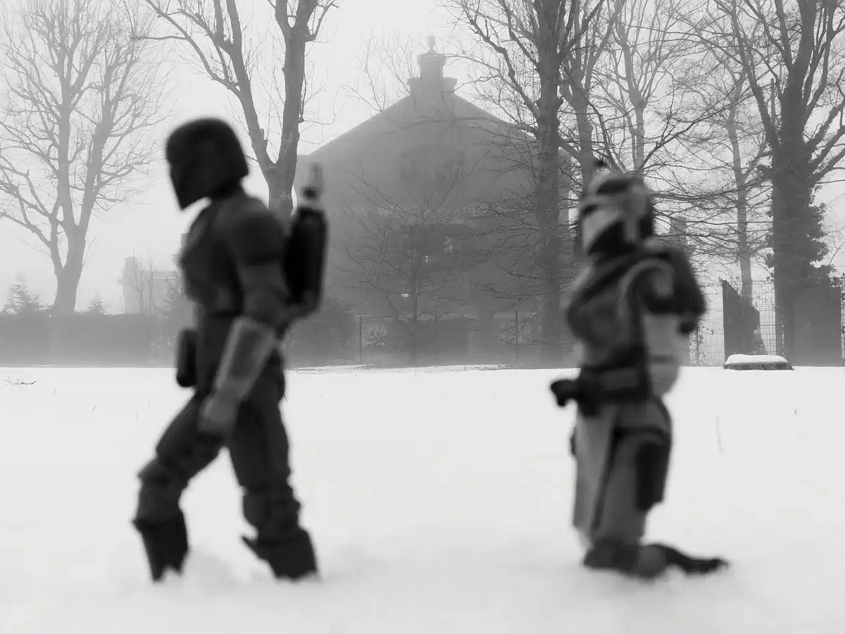 Two figures in Mandalorian armor stand in a snowy, foggy field with bare trees and a distant building. The figure on the…