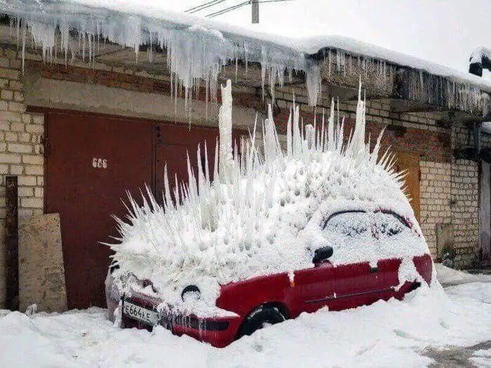 a red car is parked outside a brick building and is completely covered in snow and numerous sharp, spiky ice formations…