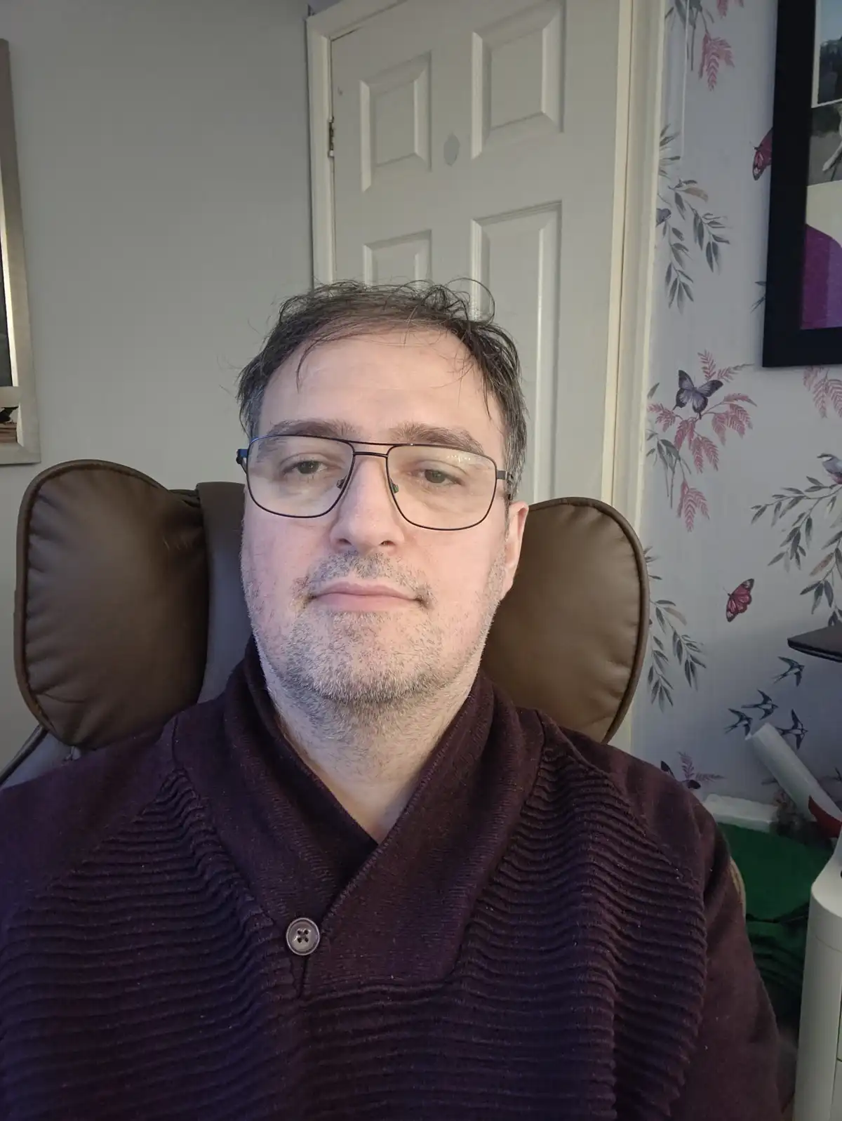 A man with glasses and a beard sits in a brown chair, looking directly at the camera with a neutral expression. He wears a…