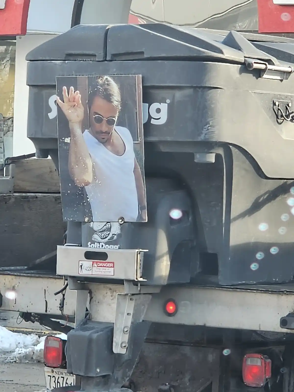 A close-up of a large, dirty black truck bed with a faded poster of a man in a white tank top and sunglasses, waving with…