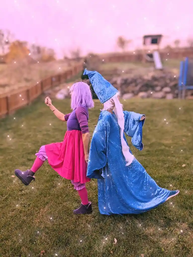 Two people in cosplay stand back-to-back on a grassy lawn, one dressed as a pink-robed wizard apprentice with purple hair…