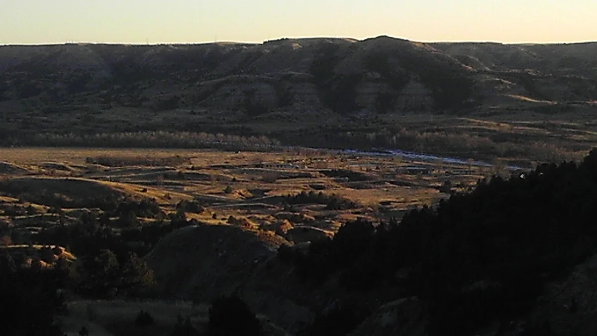A wide, sunlit landscape photo of rolling hills and valleys with sparse trees and distant snow patches under a pale sky…