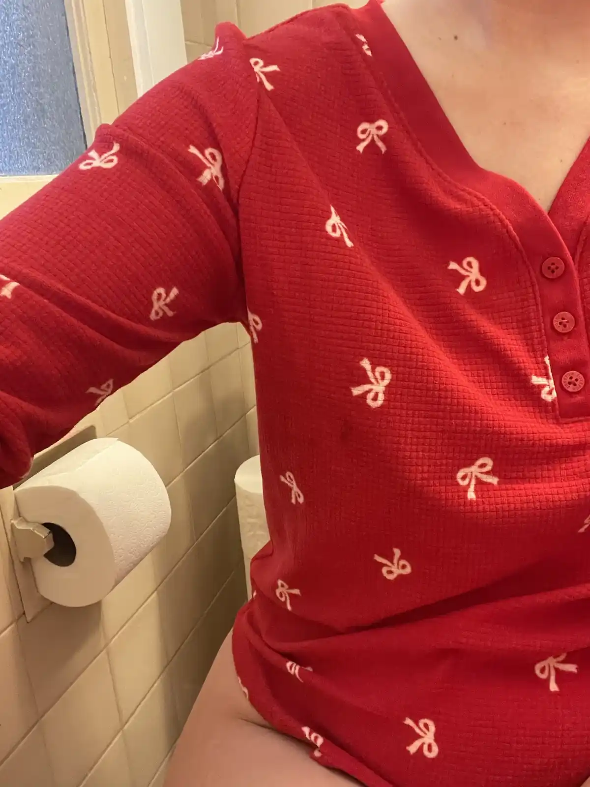 A person wearing a red long-sleeved shirt with white bow tie patterns is sitting on a toilet in a bathroom, with a toilet…