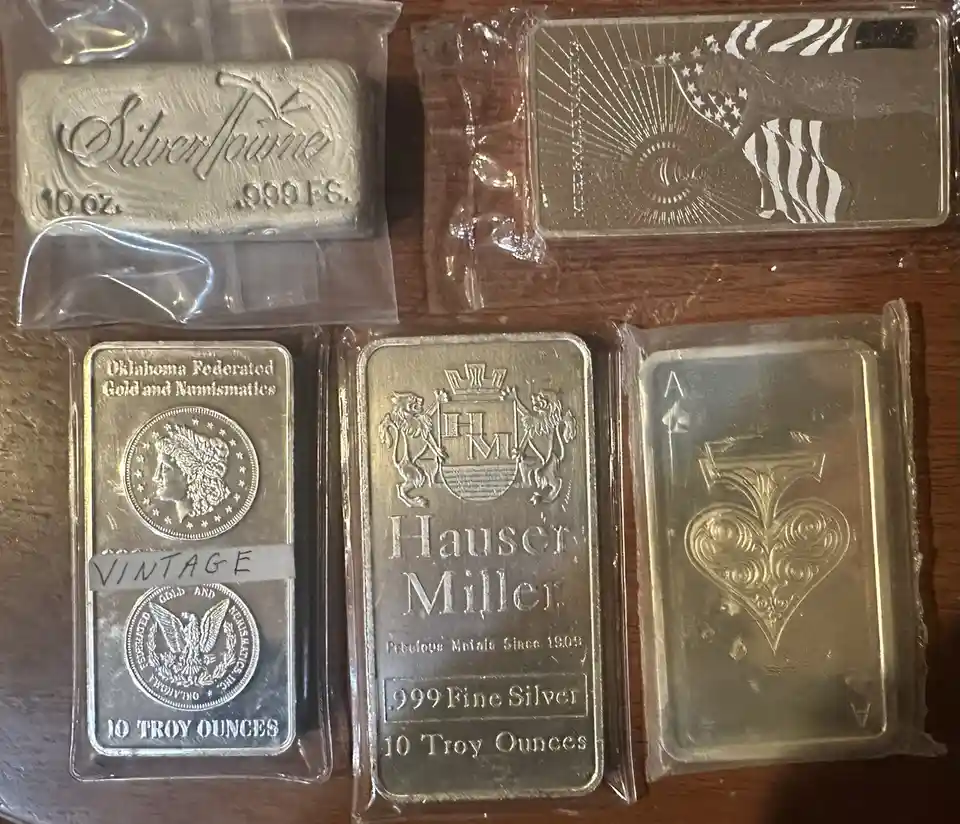 Five silver bars in plastic packaging are arranged on a wooden surface, each with different designs and markings. One bar…