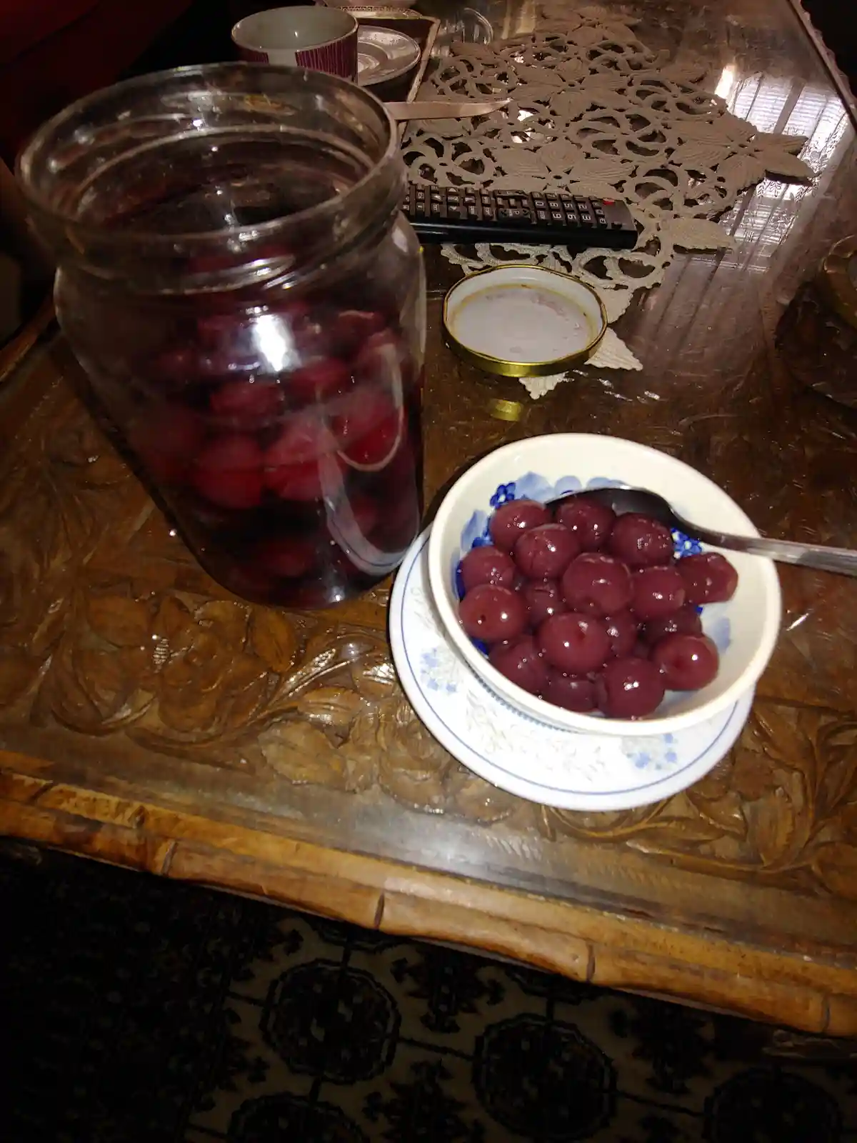 A glass jar filled with cherries in liquid sits next to a small bowl of cherries on a wooden table with a remote and lace…