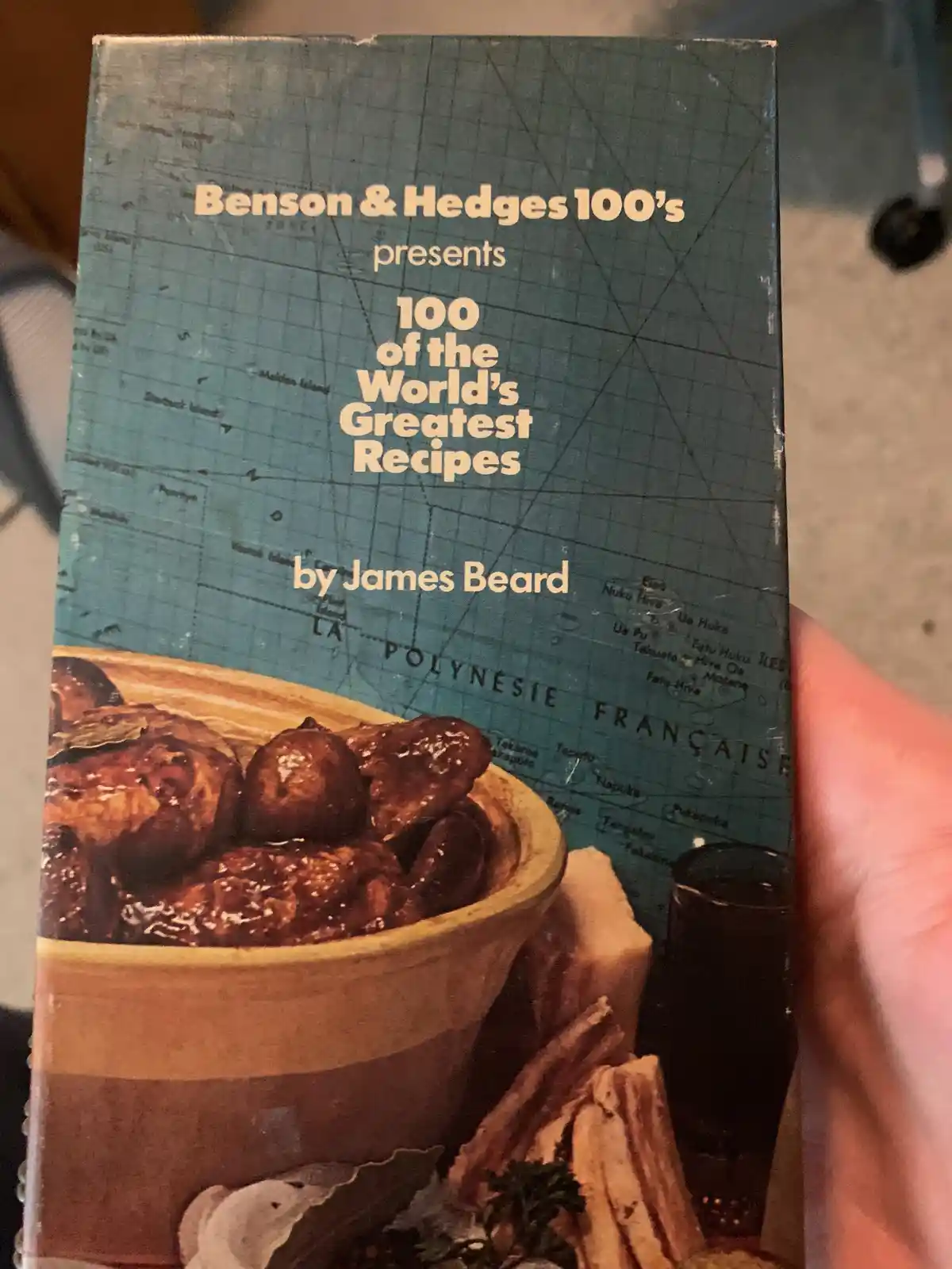 A person holds a book with a cover that reads 'Benson & Hedges 100's presents 100 of the World's Greatest Recipes by James…