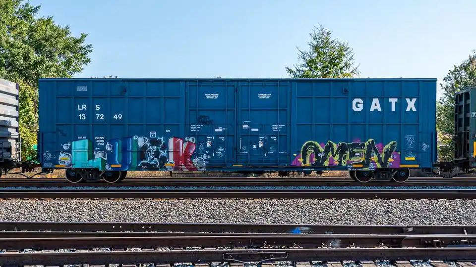 A blue GATX freight train car sits on tracks under a clear sky, covered in colorful graffiti including large stylized…