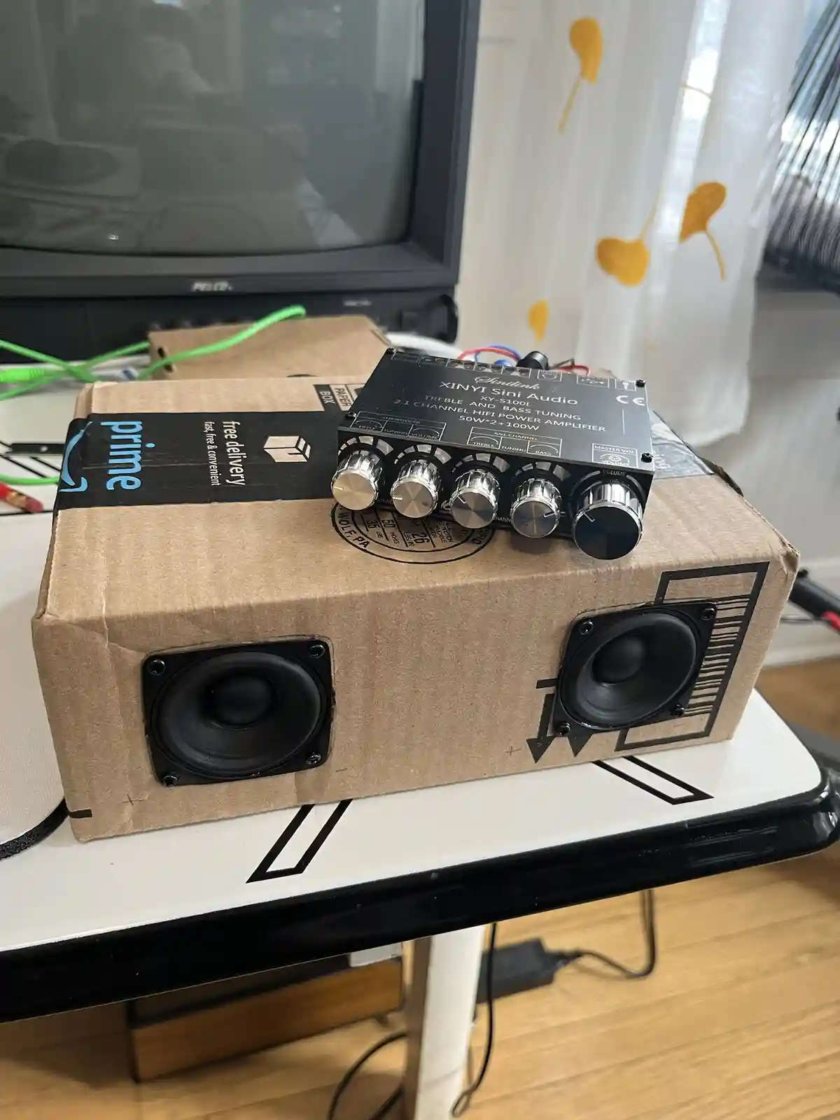 A DIY speaker setup made from a brown Amazon Prime cardboard box, with two black speaker drivers mounted on its front and a…