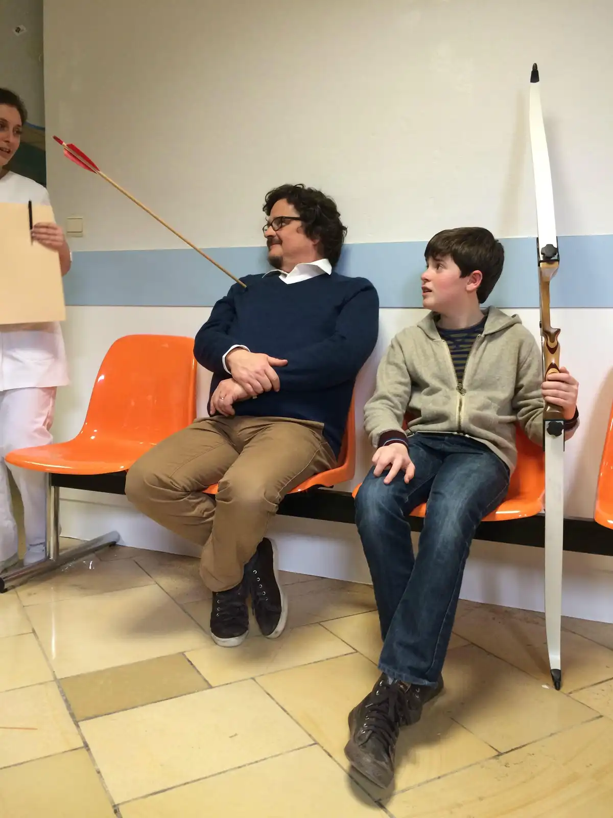 In a hospital waiting room, a man and a boy sit on orange chairs. The boy holds a bow, and an arrow is stuck in the man's…