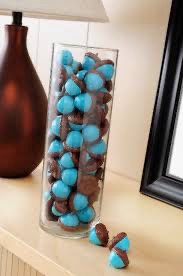 A tall glass vase filled with blue and brown chocolate candies sits on a wooden surface, with a few scattered candies…