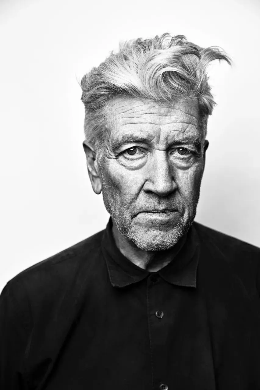 A black-and-white portrait of an older man with graying hair and a stern, weary expression, wearing a dark collared shirt…