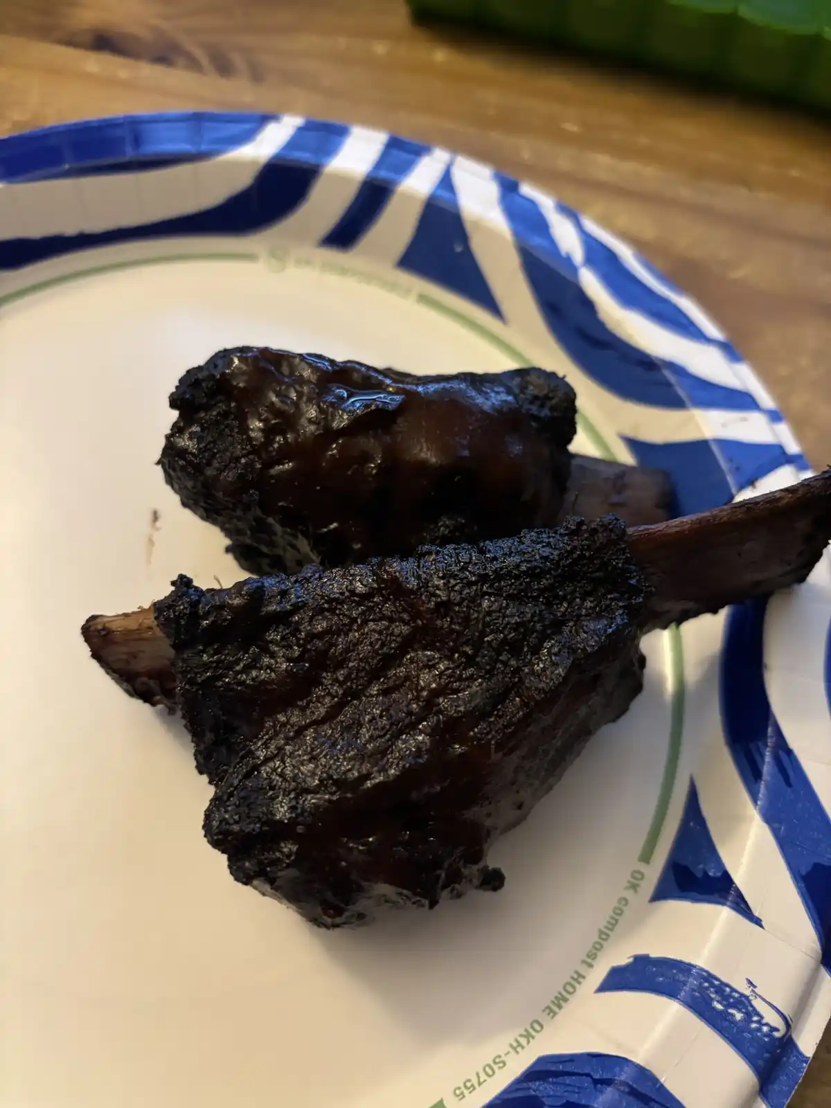 Two heavily charred, blackened pieces of meat resembling ribs sit on a blue and white striped paper plate with 'OK compost…