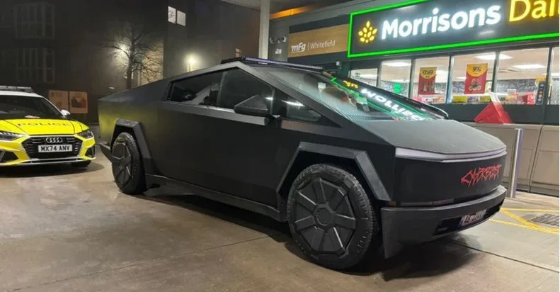a black tesla cybertruck is parked at a morrisons supermarket at night, with a yellow police car parked beside it. the…