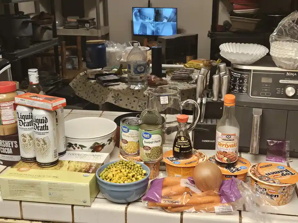 A cluttered kitchen counter is covered with various food items including canned sweet peas and corn, ramen noodles, soy…