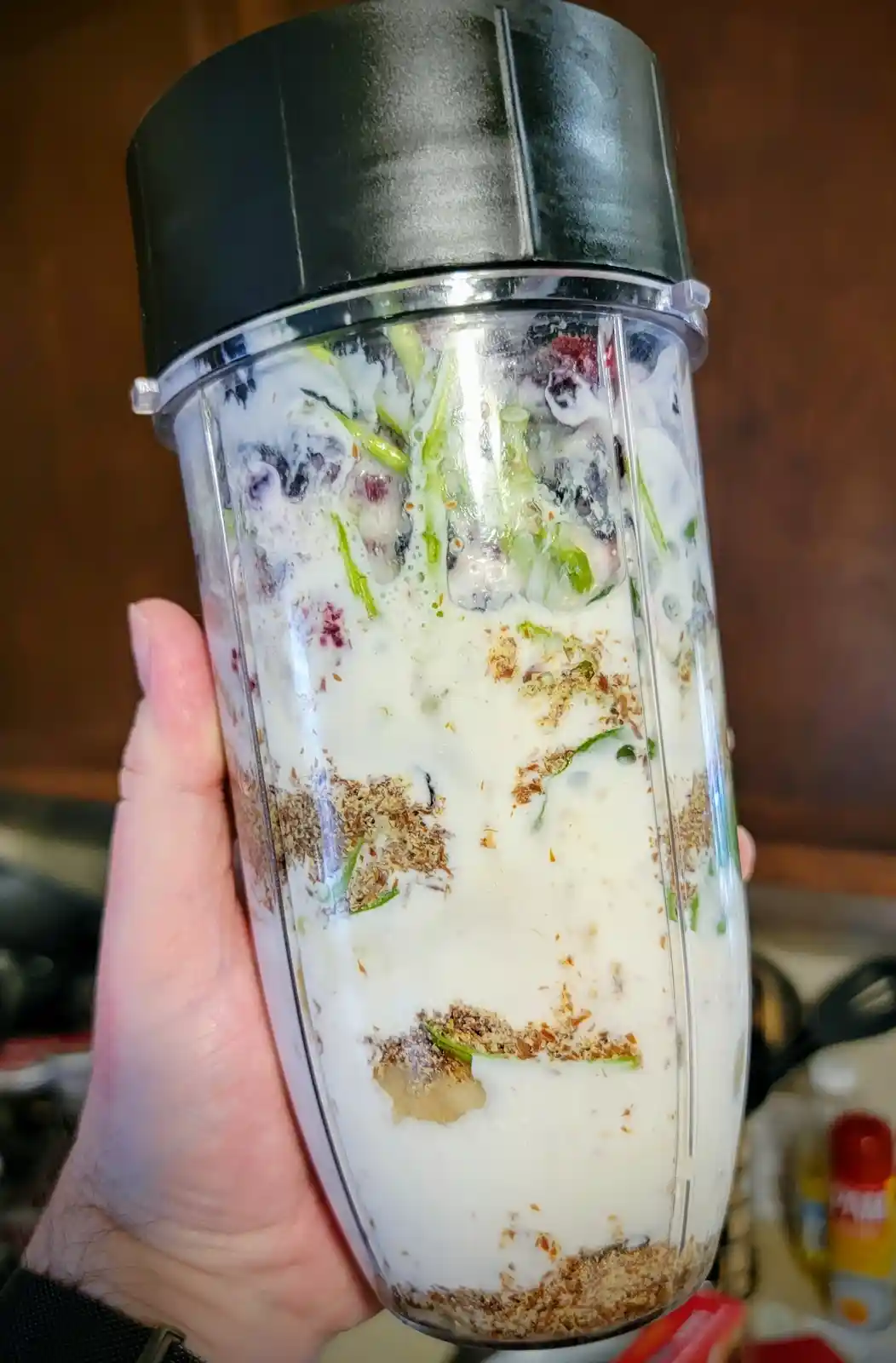 A hand holds a clear blender cup with a black lid, filled with a white liquid, frozen berries, green vegetable pieces, and…
