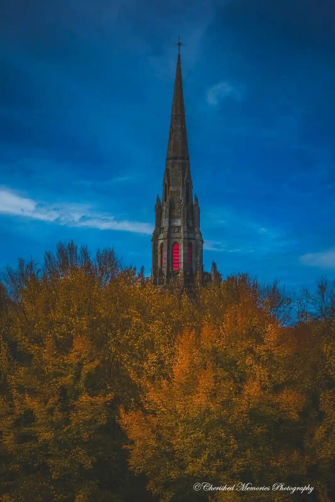 A tall, pointed church spire rises above a dense forest of orange and yellow autumn trees against a deep blue twilight sky…