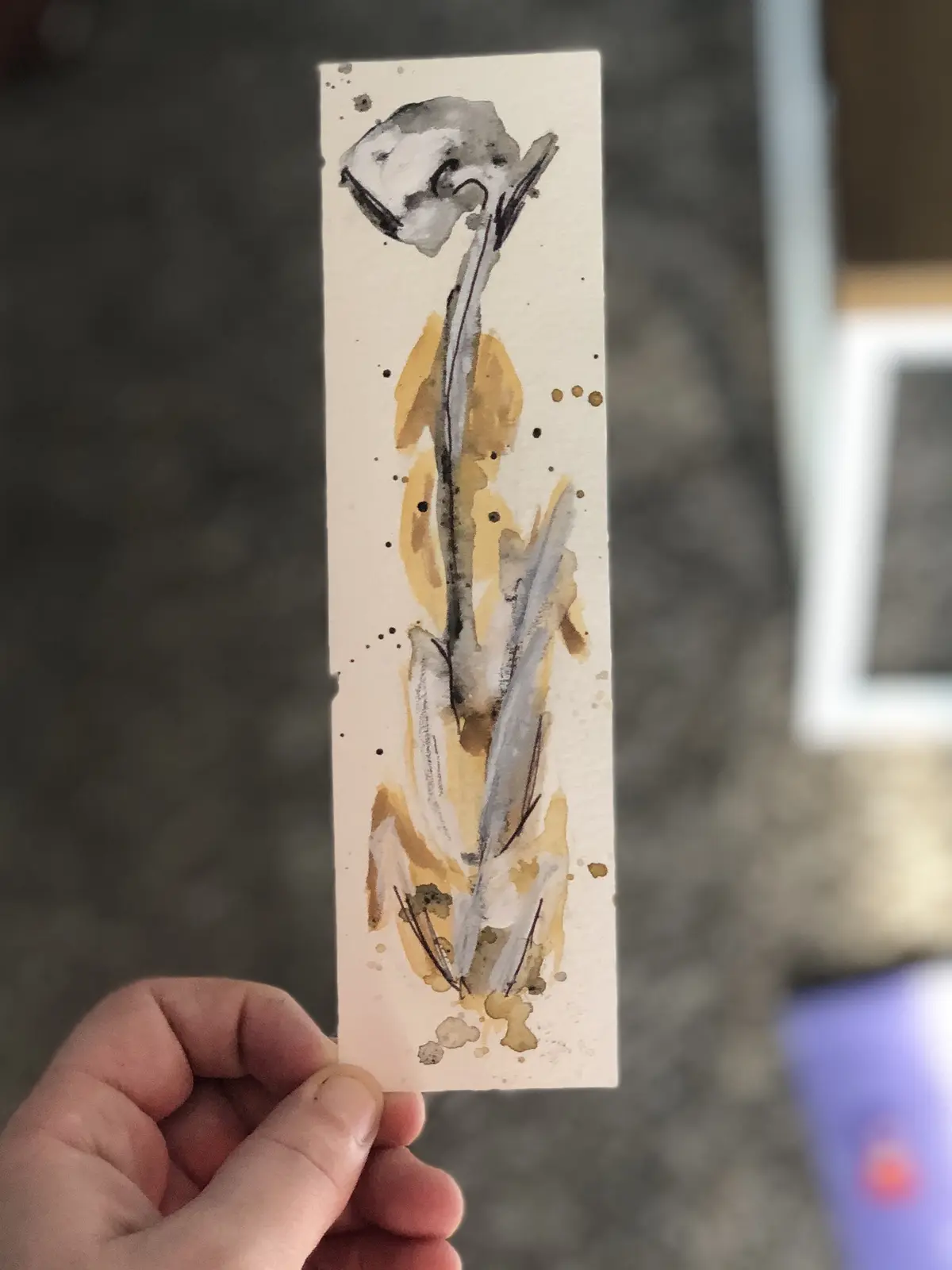 A hand holds up a vertically oriented watercolor painting of a flower on light beige paper. The flower, rendered in gray and…