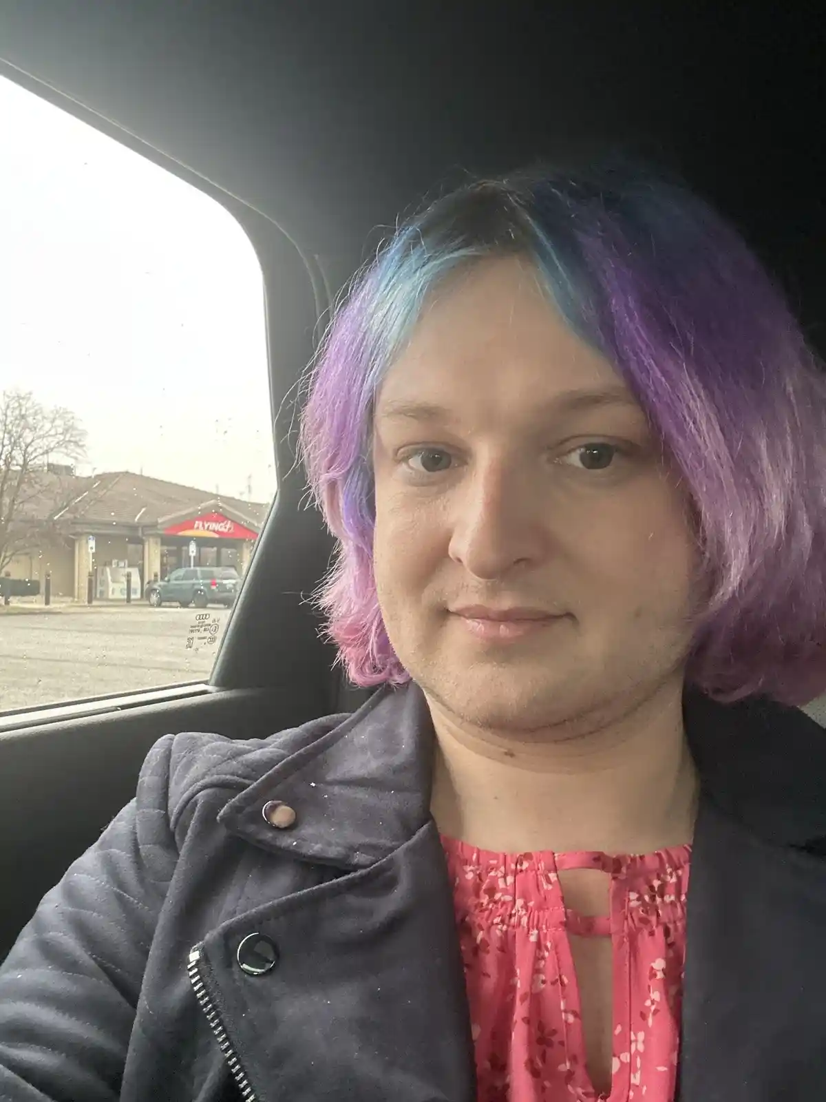 A person with short, colorful purple and blue hair sits in a car, looking at the camera with a neutral expression. They wear…