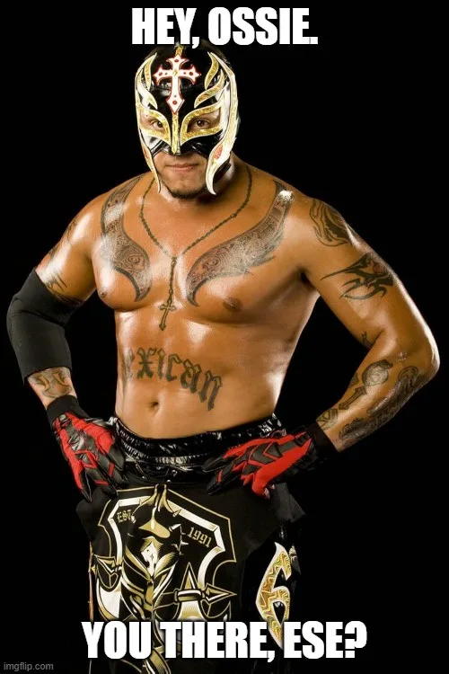 A muscular luchador in a black and gold mask and trunks stands with hands on hips against a black background. He has tattoos…