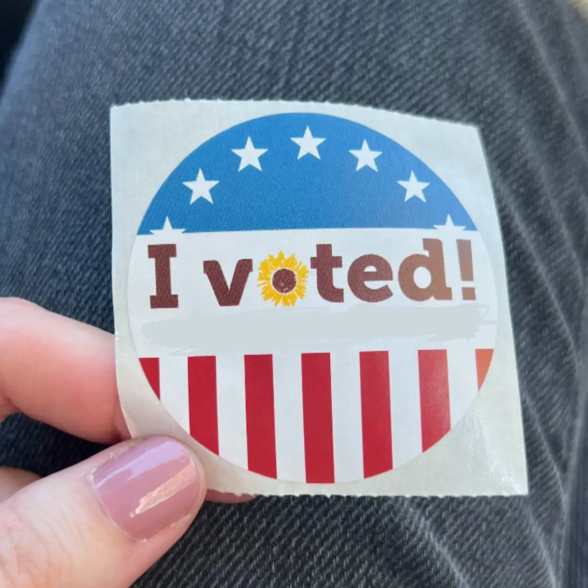 A close-up photo shows a hand holding a circular 'I voted!' sticker against a dark denim background. The sticker features a…