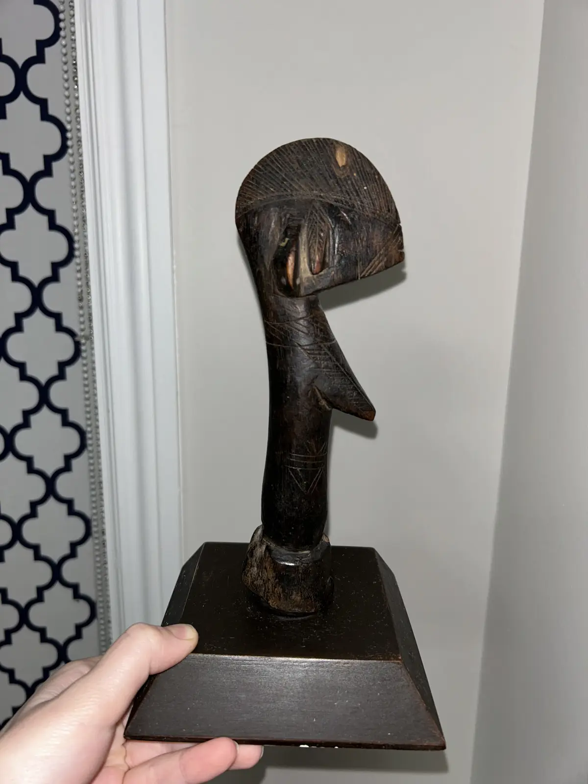 A hand holds a dark wooden sculpture on a square base, featuring a stylized, abstract human-like figure with a large head…