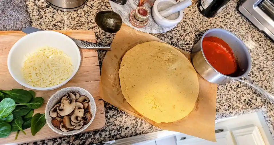 An overhead shot of a kitchen counter with pizza-making ingredients: a large tortilla on parchment paper, a pot of red…
