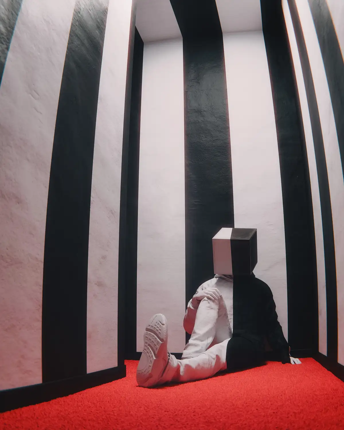 A person with a cube for a head sits on a red carpet inside a room with black and white vertical stripes. The person is…