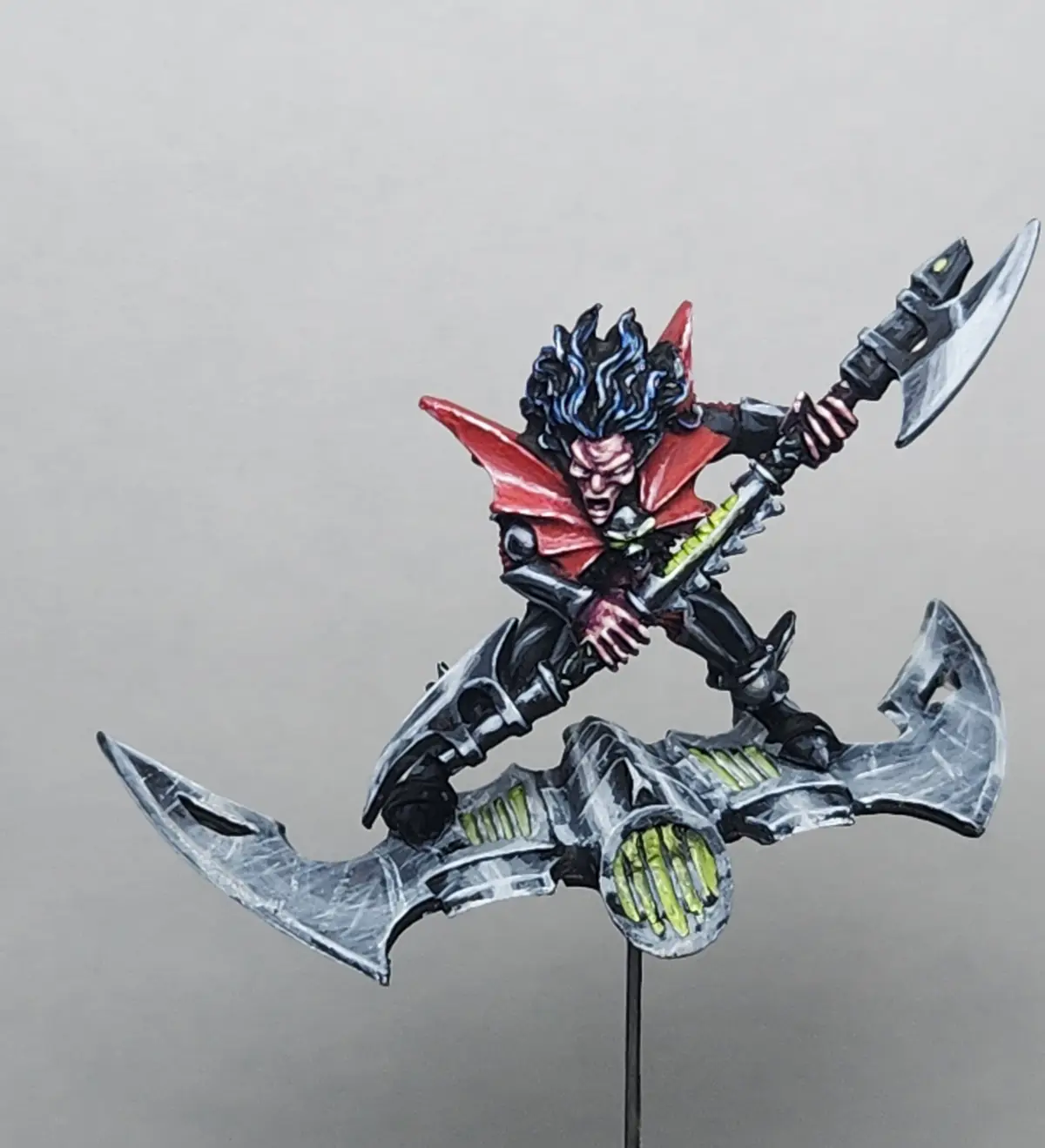 A painted miniature of a fantasy warrior with blue-black spiky hair, red cape, and black armor stands atop a large, silver…