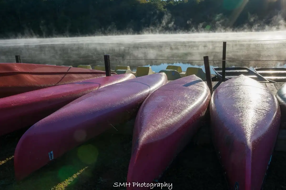 A serene photo of several red canoes lined up on a dock beside a misty lake at sunrise, with yellow paddle boats visible in…