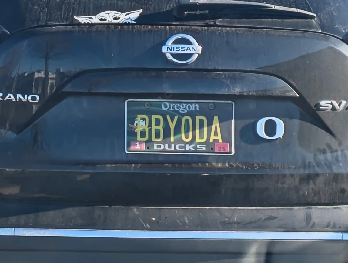 The rear of a black Nissan Rogue SUV shows an Oregon license plate with the text 'BBYODA' and 'DUCKS' below it. A small Baby…