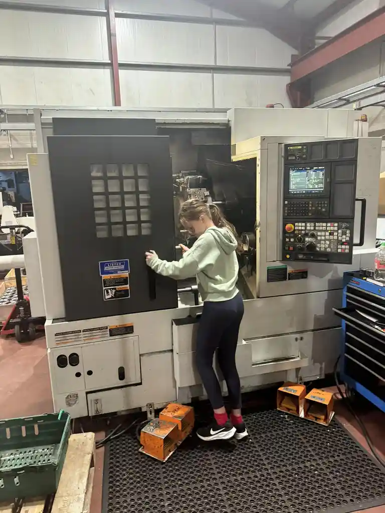 A young woman in a light green hoodie and dark leggings stands in front of a large industrial CNC machine in a factory…