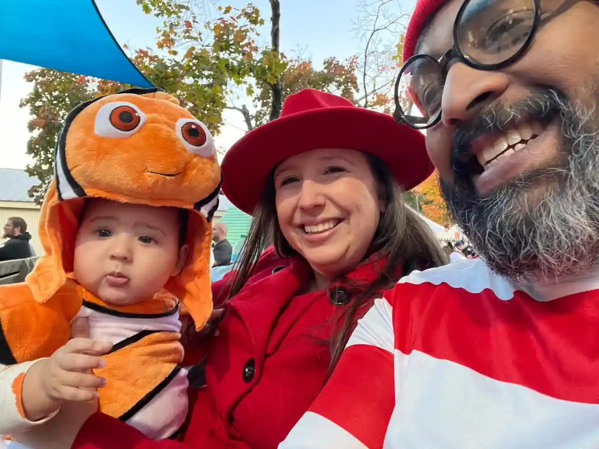 A selfie of a family of three outdoors: a baby in a clownfish costume, a woman in a red coat and hat, and a man in a…