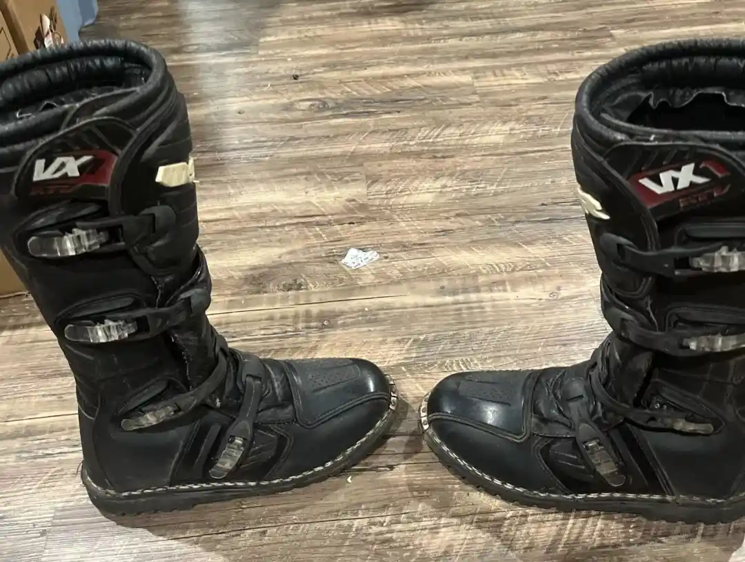 A pair of black, worn motocross-style boots with 'VX' branding sit side by side on a wooden floor. The boots show signs of…