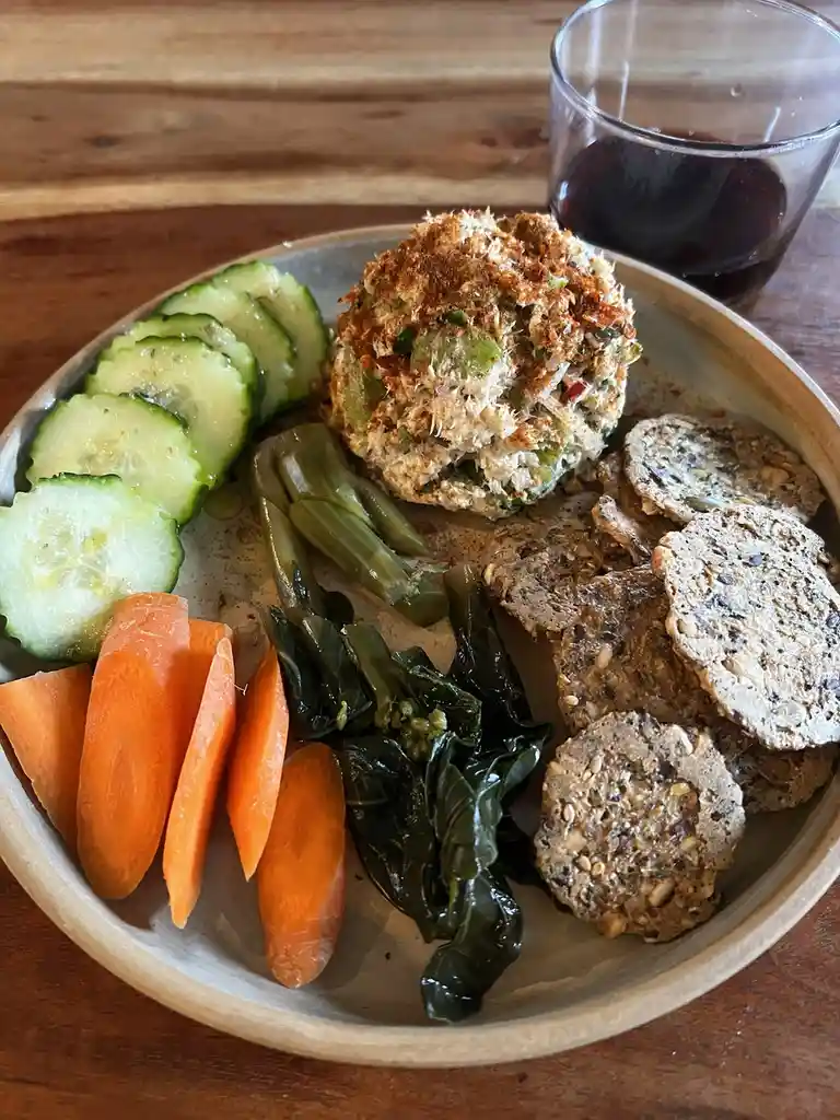 A plate of vegan food including sliced cucumbers, carrot sticks, steamed greens, seeded crackers, and a crab cake-like ball…