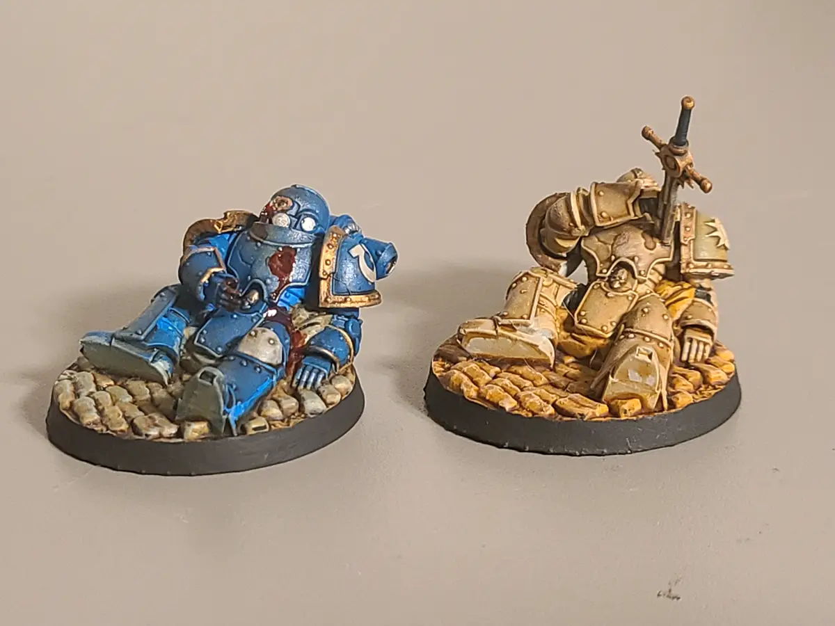 Two painted miniature space marines sit on cobblestone bases; the blue one has visible bloodstains on its chest and helmet…