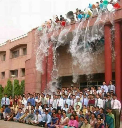 A large group of people poses for a photo in front of a building, while numerous individuals are perched on the roof holding…