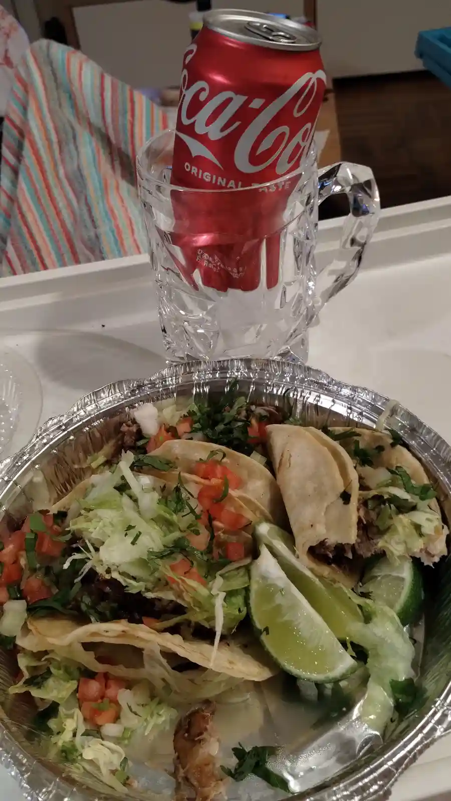 A close-up photo shows a foil tray of tacos topped with lettuce, tomatoes, and cilantro, accompanied by lime wedges. Behind…