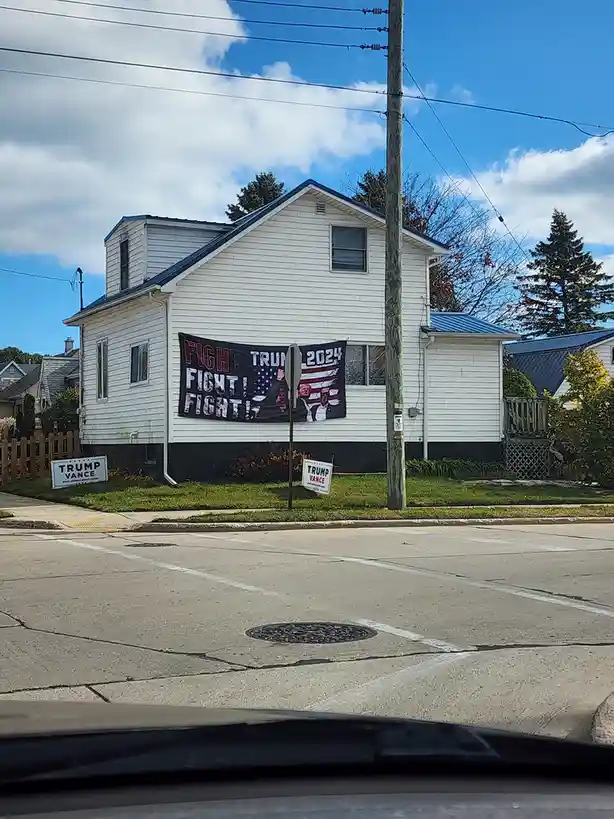 A white suburban house has a large banner on its side that reads 'FIGHT TRUMP 2024 FIGHT! FIGHT!' with an American flag…