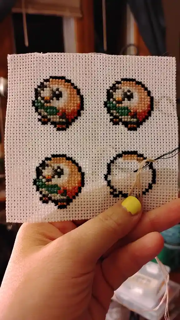 A hand holds a piece of white cross-stitch fabric with four pixelated owl characters stitched in a grid. Three owls are…