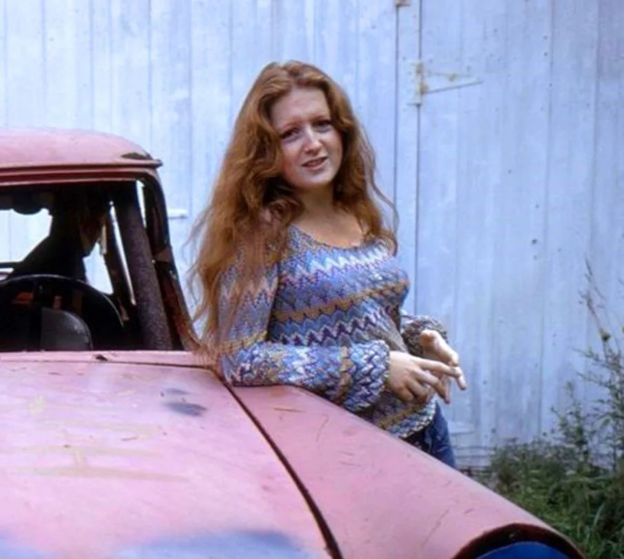 a woman with long red hair wearing a patterned sweater leans against the hood of a rusty, old red car. Behind her is a…