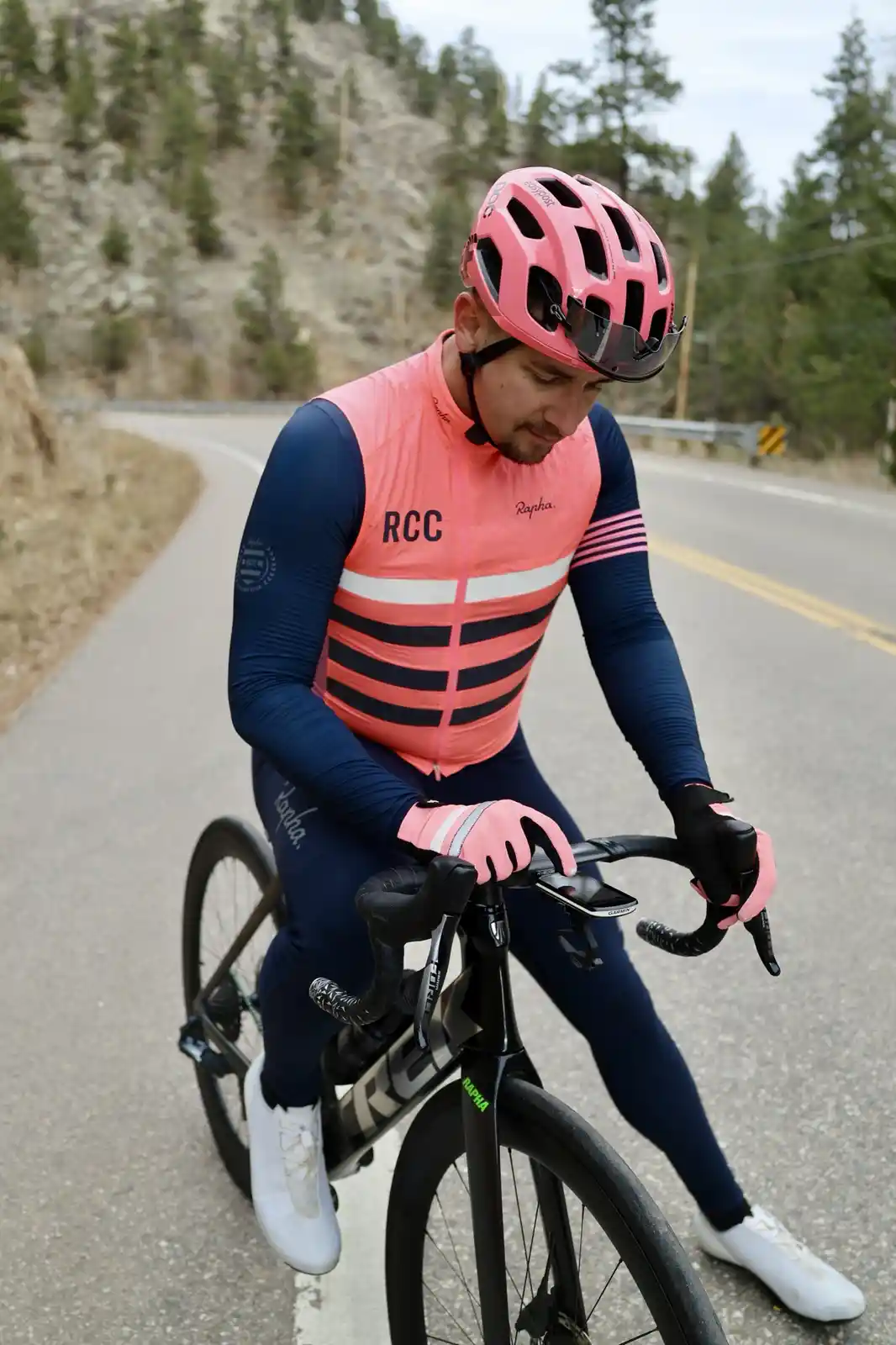 A cyclist in a pink and navy Rapha cycling kit with a matching pink helmet is stopped on a winding road, looking down at a…