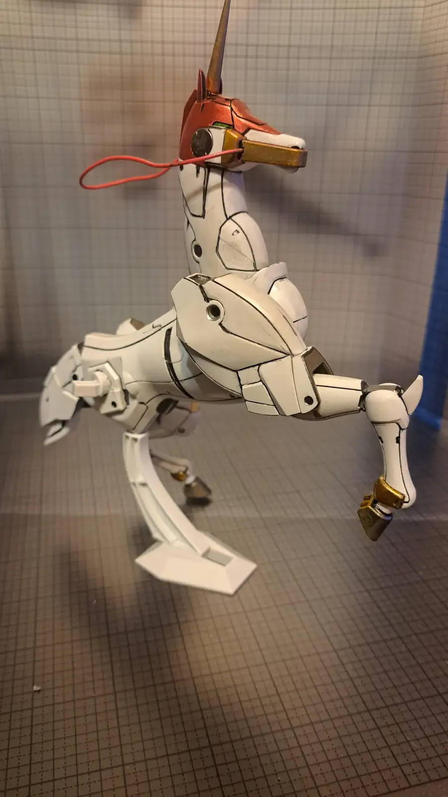A white, mechanical unicorn model with a red string tied around its mouth, standing on a display stand against a…