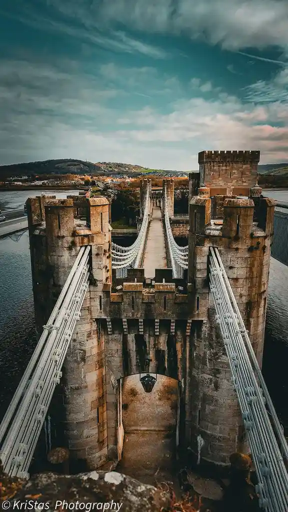 A high-angle photograph of the Conwy Suspension Bridge in Wales, showing its stone towers and suspension cables against a…