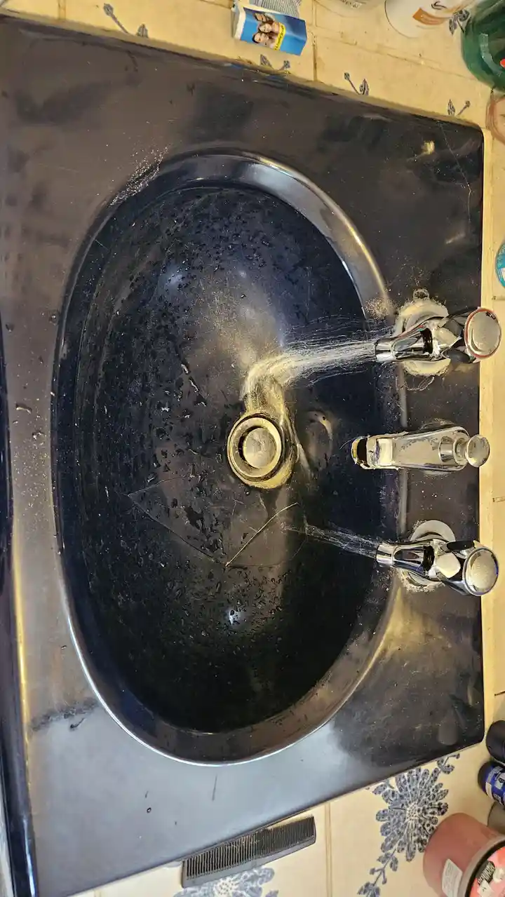 A top-down photo of a very dirty, black sink with water running from both faucets, creating a messy, soapy stream. The sink…