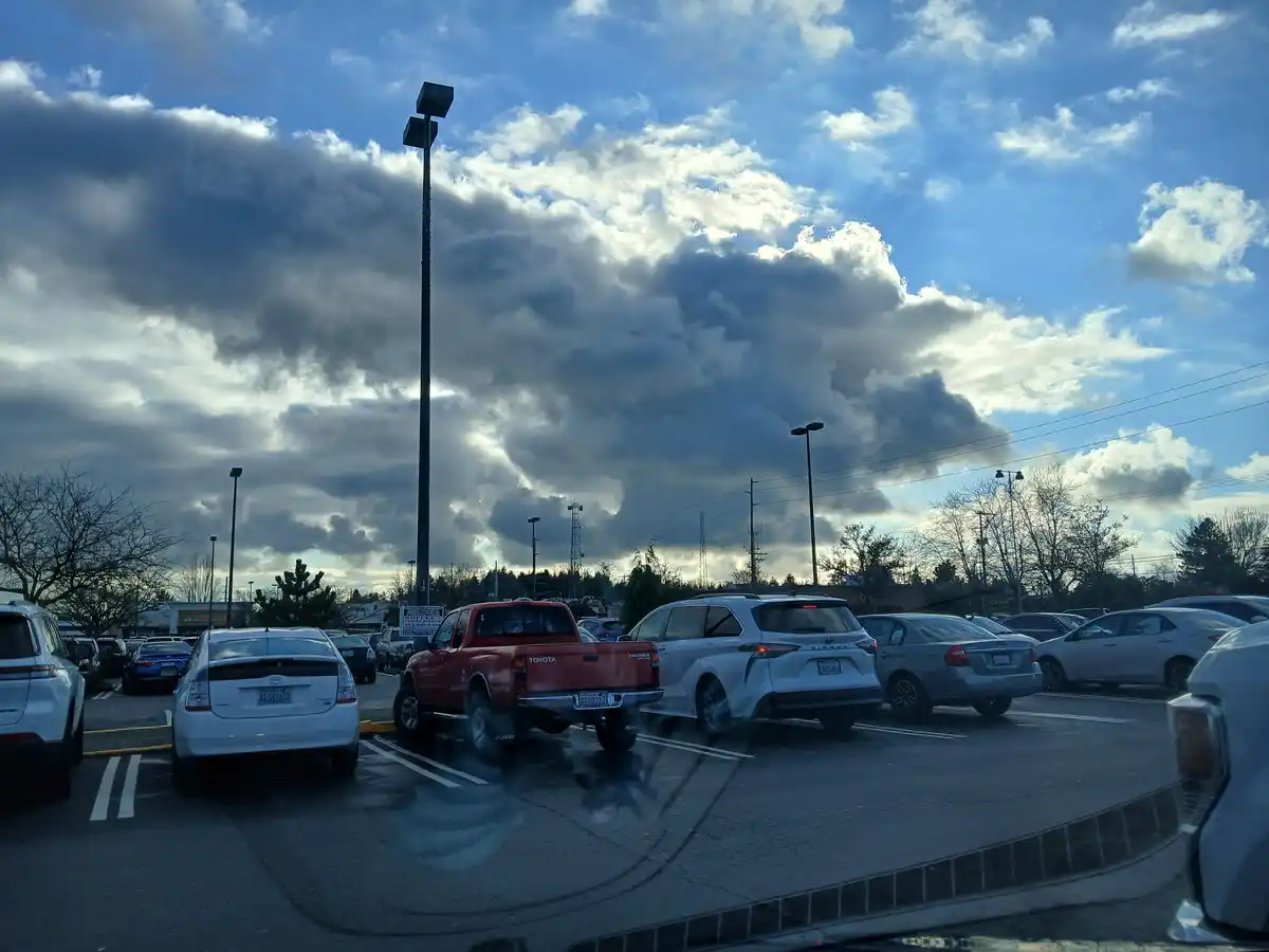 A photo taken from inside a car in a crowded parking lot shows a dramatic, dark, and heavy cloud formation looming over the…