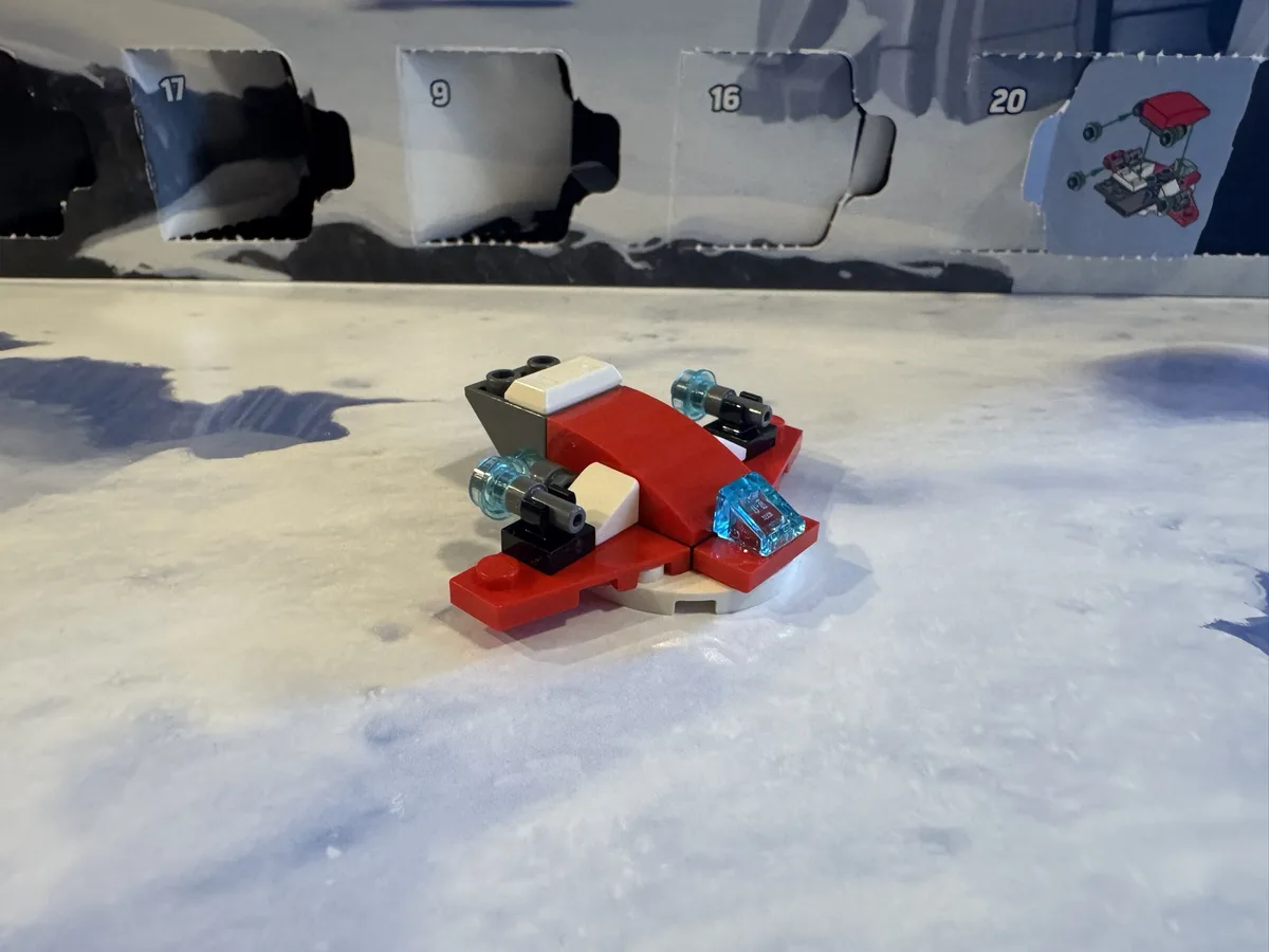 A small, partially assembled red, white, and black LEGO spaceship model sits on a white, textured surface, with the torn…