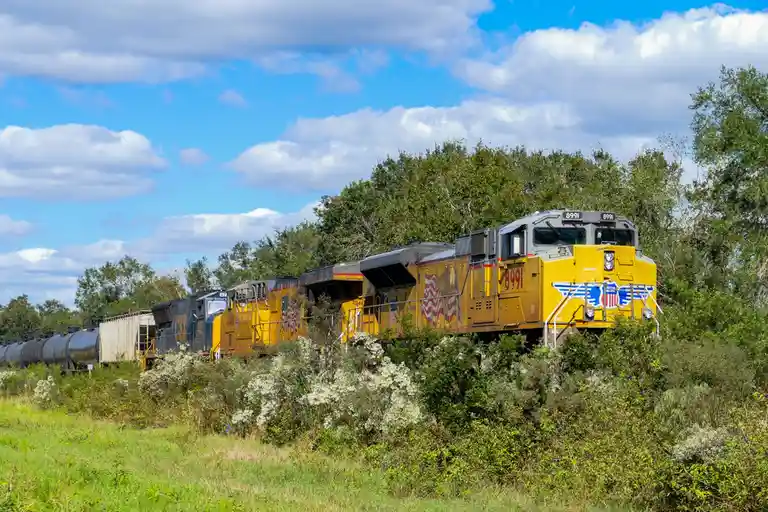 A yellow Union Pacific locomotive, number 8991, leads a freight train through greenery under a partly cloudy sky. The train…