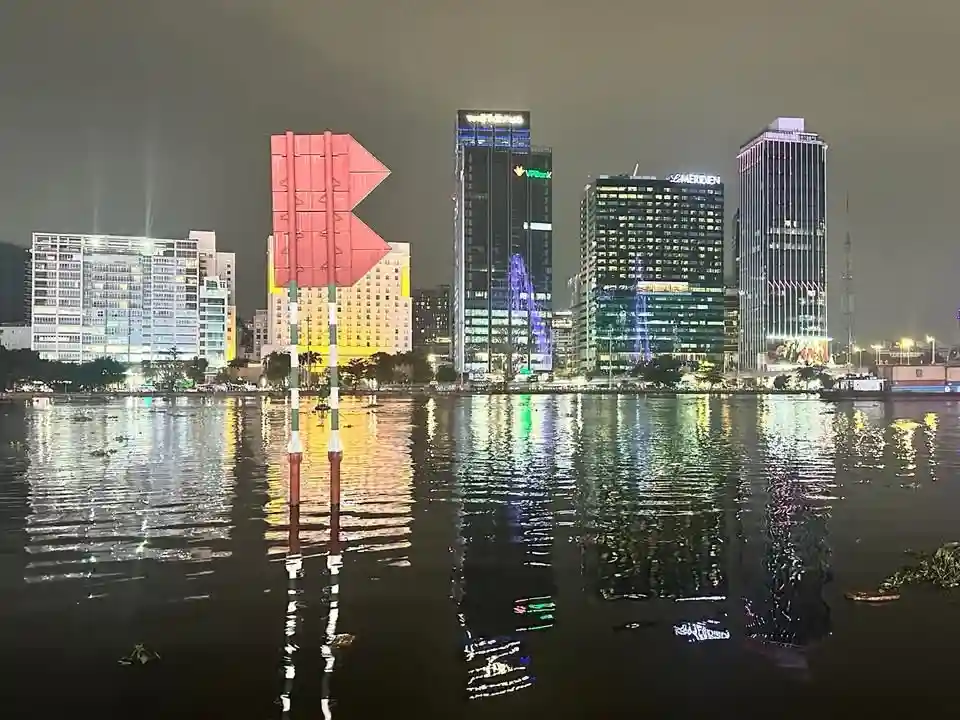 a nighttime photo shows a large red arrow-shaped buoy floating in water, with a brightly lit city skyline reflected in the…